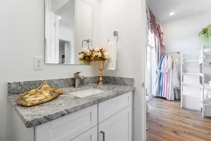 Bright modern bathroom vanity with gray granite countertop, white cabinets, gold vase of flowers and decorative tray, opening to a sunlit dressing nook with pastel dresses on a rack and white shelving on hardwood floor