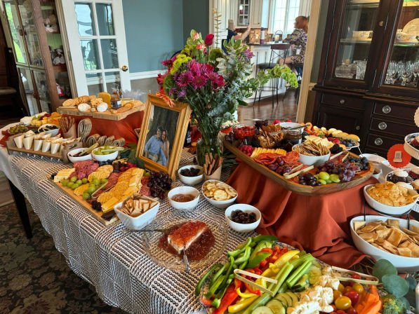 Indoor party charcuterie spread on a dining table with colorful flower centerpiece and framed photo, featuring assorted cheeses, cured meats, crackers, fruits, dips and vegetable platters.