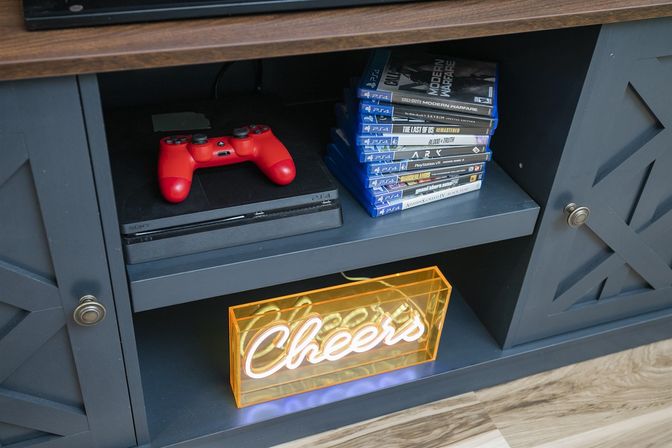 Living room media console shelf with a black PS4 and red controller, stacked PS4 game cases, and a glowing yellow-orange "Cheers" neon sign.