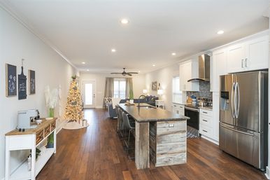 Bright open-concept kitchen and living area with dark hardwood floors, rustic wood island with bar stools, stainless-steel refrigerator, white cabinets, recessed lighting, and a decorated lit Christmas tree by the back door.