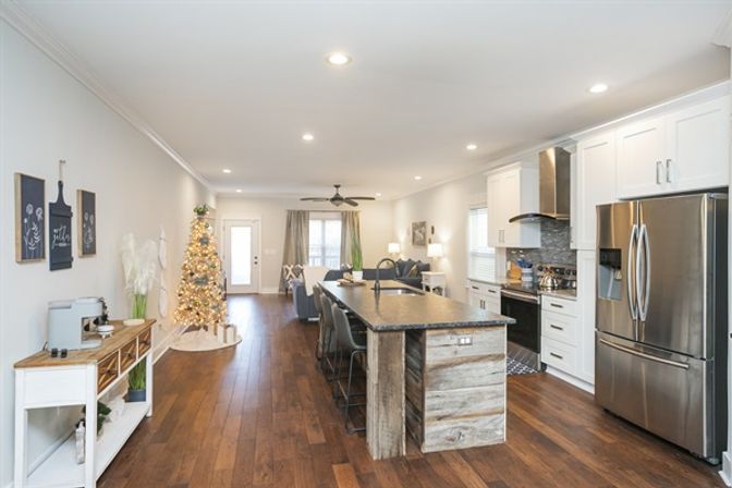 Bright open-concept kitchen and living area with dark hardwood floors, rustic wood island with bar stools, stainless-steel refrigerator, white cabinets, recessed lighting, and a decorated lit Christmas tree by the back door.