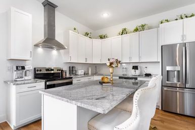 Bright modern white kitchen with a large gray granite island, tufted white bar stools, stainless steel refrigerator and range, white shaker cabinets and decorative greenery