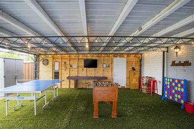 Covered backyard game room with artificial turf, string lights and Nashville-themed sign, featuring a ping-pong table, foosball, shuffleboard, dartboard, wall TV and oversized Connect Four