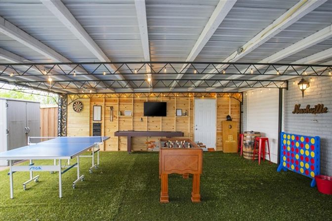 Covered backyard game room with artificial turf, string lights and Nashville-themed sign, featuring a ping-pong table, foosball, shuffleboard, dartboard, wall TV and oversized Connect Four