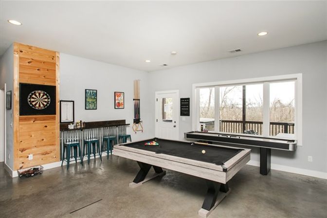 Bright modern home game room with black-felt pool table, shuffleboard by large window overlooking deck, dartboard on wood panel, metal bar stools and concrete floor.