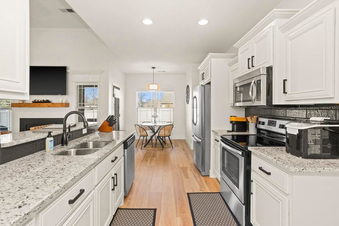 Bright modern open-plan kitchen with white cabinets, granite countertops, stainless steel appliances, undermount sink, hardwood floors and a sunny breakfast nook with a round table by a large window.