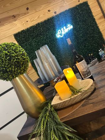Cozy rustic bar vignette: bottle of red wine, flameless candles on a wooden tray, stacked plastic cups, gold vase with faux topiary, and a green boxwood wall with a glowing neon "Welcome" sign.