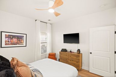 Cozy modern bedroom with crisp white walls and wood floor, bed with neutral pillows, wooden dresser topped with books and decor, wall-mounted flat-screen TV, orange travel trunks by a curtained window, and a three-blade wood ceiling fan.