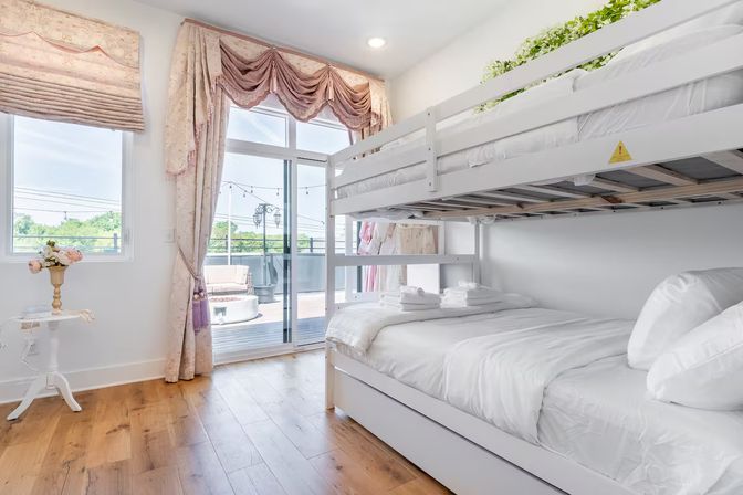 Bright vacation-rental bedroom with white bunk beds and crisp linens, hardwood floors, pink floral curtains, small side table with flowers, and sliding glass doors opening to a sunny balcony with outdoor seating and string lights.