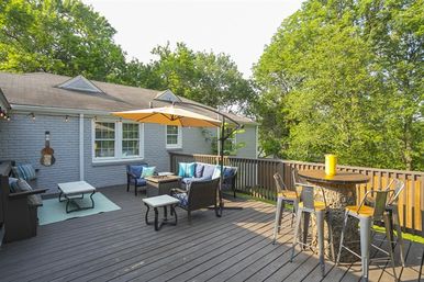 Sunny suburban backyard deck with gray brick house, wicker seating and blue cushions under a yellow patio umbrella, fire-pit coffee table, and a high round bar table with metal stools overlooking green trees.