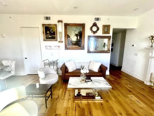 Sunlit cozy-chic living room with oak hardwood floors, brown leather sofa topped with white pillows, marble-top coffee table with vase of flowers, gallery wall of framed paintings and mirrors, and mixed modern-vintage seating.