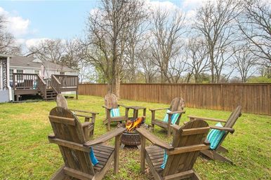 Cozy fenced suburban backyard with a wooden deck, six Adirondack chairs with turquoise-striped cushions arranged around a lit fire pit on a grassy lawn under bare trees.
