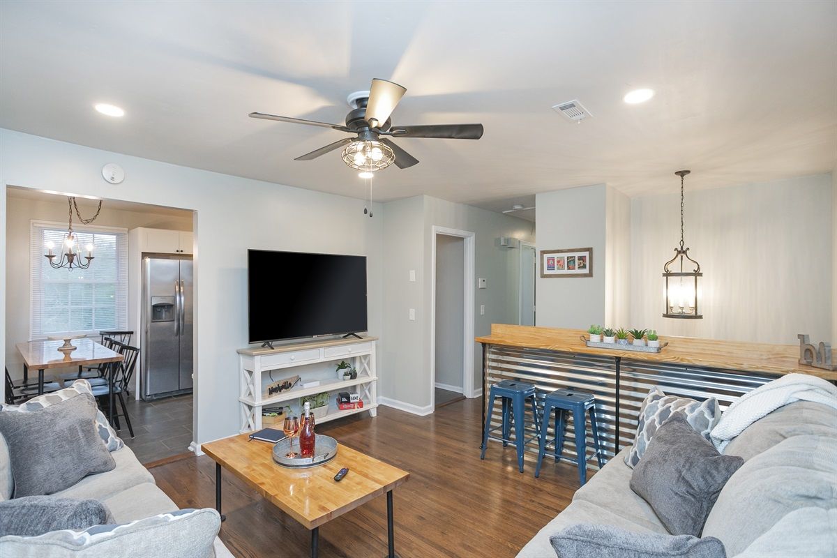 Cozy open-concept living room and kitchen in a modern home with hardwood floors, gray sofas, ceiling fan, wall TV, and corrugated-metal breakfast bar with blue stools.