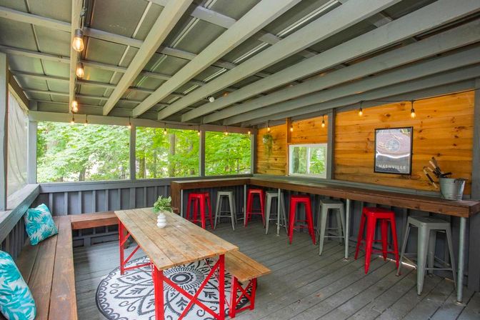 Screened-in rustic porch patio with long wooden bar, red metal stools, picnic table and bench, string lights, and leafy woodland view.