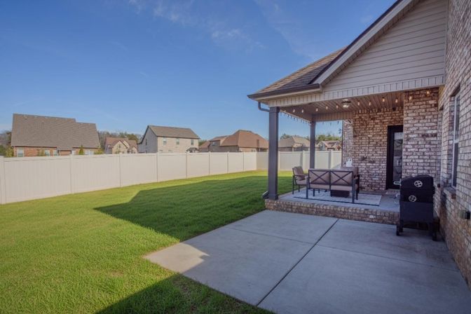 Suburban backyard with green lawn, white vinyl privacy fence, covered brick patio featuring outdoor seating and a grill under a clear blue sky.