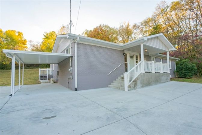 One-story light-gray brick ranch house with a covered carport, wide concrete driveway, front steps and white-railed porch, surrounded by autumn trees in a suburban residential lot.