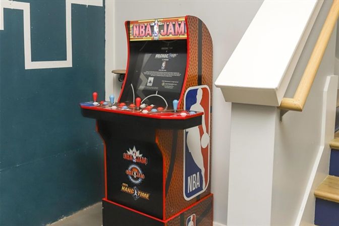 NBA Jam arcade machine with red-and-black cabinet and colorful joysticks tucked beside an indoor staircase
