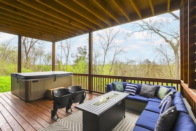Cozy covered wooden backyard deck with a covered hot tub, two black stadium-style chairs, navy blue outdoor sectional with patterned pillows and a fire‑pit table overlooking a wooded yard.