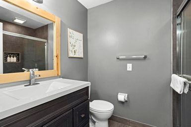 Contemporary gray bathroom with dark wood vanity and white rectangular sink, natural wood-framed mirror, chrome faucet, white toilet, glass shower with towel, and botanical wall art — clean, modern interior.