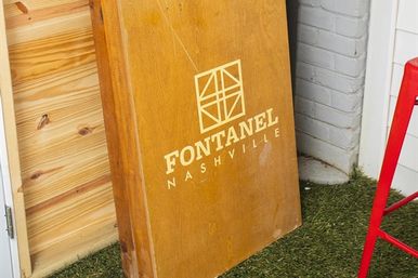 Wooden display sign with geometric logo and lettering leaning against a painted brick wall on artificial turf, red metal stool nearby — Nashville outdoor scene.