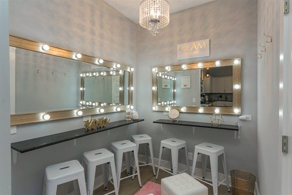 Glam makeup vanity room with two Hollywood-style lighted mirrors, crystal chandelier, floating black counters, five white metal stools and a pink rug in a cozy gray-walled space