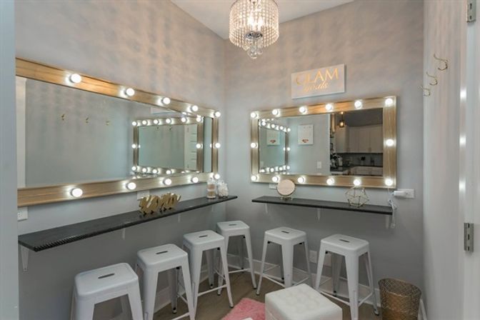 Glam makeup vanity room with two Hollywood-style lighted mirrors, crystal chandelier, floating black counters, five white metal stools and a pink rug in a cozy gray-walled space