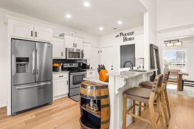 Bright modern open-concept kitchen with white cabinets, stainless steel fridge and stove, granite breakfast bar with wooden bar stools, repurposed oak barrel shelf, light hardwood floors and recessed lighting.