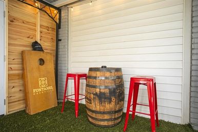 Covered patio seating featuring a wooden whiskey barrel used as a table flanked by two red metal bar stools and a cornhole game board leaning against a wood-paneled wall on faux grass