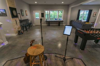 Basement game room and entertainment space with small stage area—stool and guitar, microphone and tablet on stands—concrete floor, foosball table, shuffleboard by large windows, dartboard and bar stools along a gray wall.