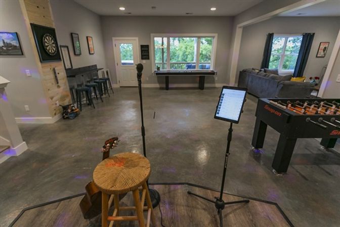 Basement game room and entertainment space with small stage area—stool and guitar, microphone and tablet on stands—concrete floor, foosball table, shuffleboard by large windows, dartboard and bar stools along a gray wall.