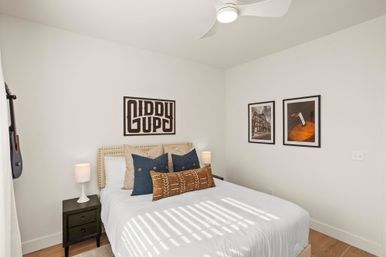 Bright modern bedroom with white walls and wood floors, a bed with beige nailhead headboard layered with navy and patterned pillows, black nightstands with white lamps, framed wall art, a ceiling fan, a hanging acoustic guitar, and sunlight casting striped shadows on white bedding.