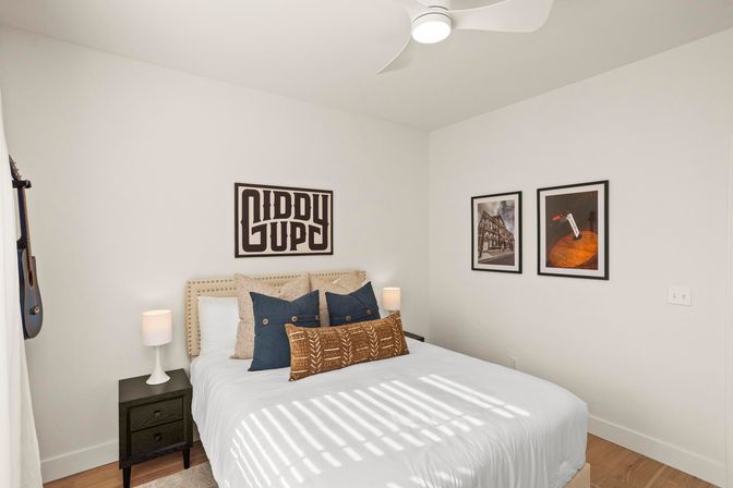 Bright modern bedroom with white walls and wood floors, a bed with beige nailhead headboard layered with navy and patterned pillows, black nightstands with white lamps, framed wall art, a ceiling fan, a hanging acoustic guitar, and sunlight casting striped shadows on white bedding.