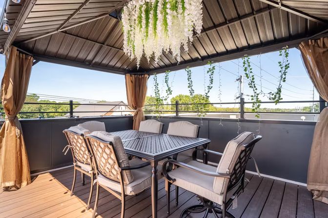 Cozy rooftop gazebo with metal roof and draped curtains, outdoor dining table and cushioned chairs, hanging white wisteria and string lights, railing overlooking treetops and suburban rooftops