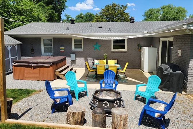 Sunny suburban backyard patio with a covered hot tub, yellow outdoor dining set under a shade sail, bright blue and teal Adirondack chairs arranged around a metal fire pit with star cutouts and tree-stump stools on gravel.