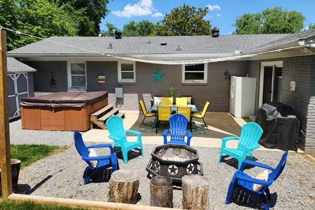 Sunny suburban backyard patio with a covered hot tub, yellow outdoor dining set under a shade sail, bright blue and teal Adirondack chairs arranged around a metal fire pit with star cutouts and tree-stump stools on gravel.