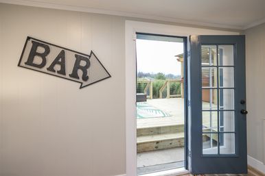 Interior room with metal "BAR" arrow sign on the wall and an open blue French door leading to a sunlit wooden deck and backyard greenery.