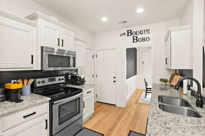 Bright modern white kitchen with stainless steel stove and microwave, speckled granite countertops, black tile backsplash, farmhouse sink, light hardwood floors, and wall decal reading "The Bougie Boro" leading to a small dining nook.