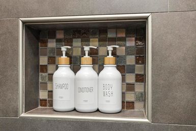 Three white pump bottles labeled Shampoo, Conditioner and Body Wash neatly arranged in a mosaic-tiled shower niche in a modern gray bathroom