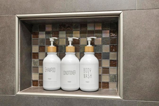 Three white pump bottles labeled Shampoo, Conditioner and Body Wash neatly arranged in a mosaic-tiled shower niche in a modern gray bathroom