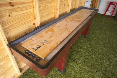 Indoor shuffleboard table with a worn wooden playing surface and four black pucks against a pine-paneled wall on artificial turf in a game room