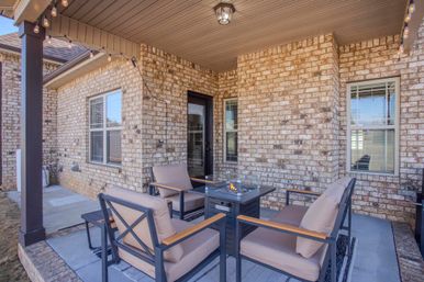 Covered brick backyard patio at a suburban home with four cushioned armchairs arranged around a lit square fire pit table, overhead string lights, windows and a glass door — cozy outdoor seating area.