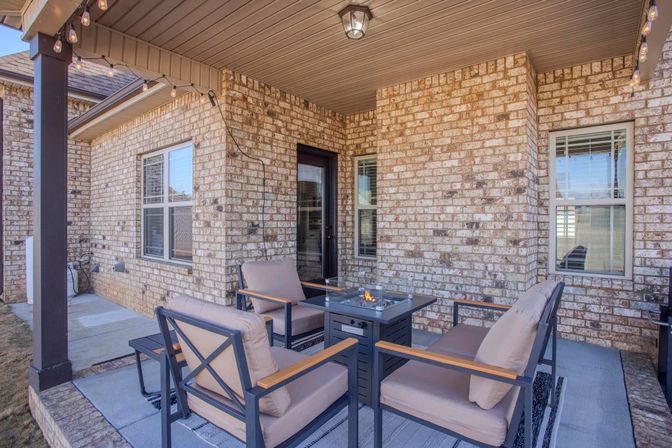 Covered brick backyard patio at a suburban home with four cushioned armchairs arranged around a lit square fire pit table, overhead string lights, windows and a glass door — cozy outdoor seating area.