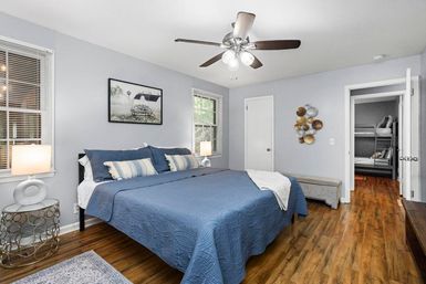 Modern master bedroom with blue quilted king bed, hardwood floors, ceiling fan, bedside lamps and an open doorway revealing bunk beds in the adjoining room.