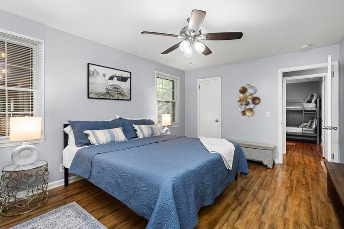 Modern master bedroom with blue quilted king bed, hardwood floors, ceiling fan, bedside lamps and an open doorway revealing bunk beds in the adjoining room.