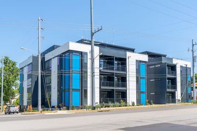 Modern three-story apartment building with bright blue vertical accent panels and balconies on a sunny urban street with utility poles