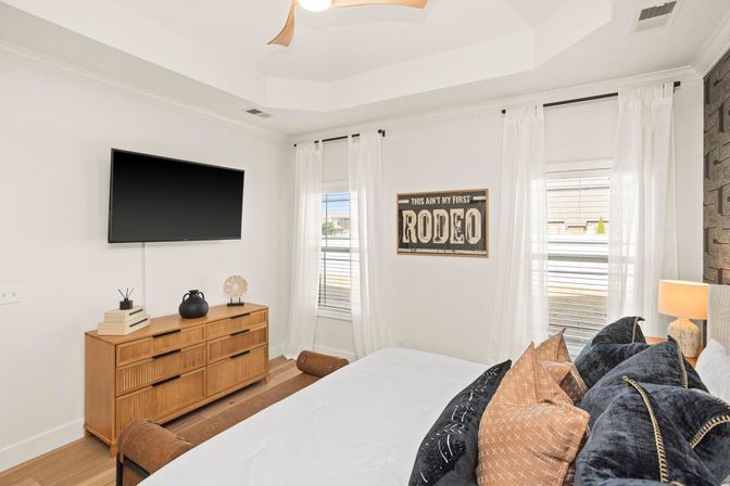 Bright modern bedroom with wall-mounted flat-screen TV, wooden dresser, airy white curtains, a 'This Ain't My First Rodeo' wall sign, and a bed layered with patterned throw pillows