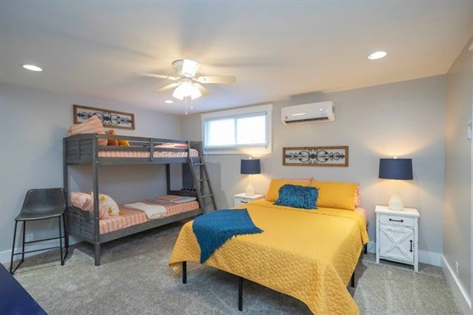 Bright family bedroom with a gray bunk bed and a queen bed dressed in a yellow quilt and blue accents, white nightstands with navy lamps, ceiling fan, wall-mounted air conditioner, and carpeted floor.
