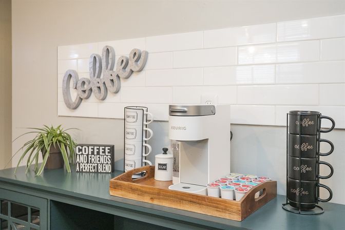 Cozy home coffee bar with a white Keurig on a wooden tray, colorful coffee pods, stacked black mugs, a sugar jar, potted plant and decorative 'Coffee' sign on a tiled backsplash.