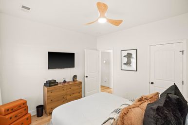 Bright contemporary bedroom with white walls, light oak floors, wooden ceiling fan, wall-mounted flat-screen TV above a mid-century wooden dresser, stacked orange suitcases, framed hat print, and a bed dressed with rust and black pillows.