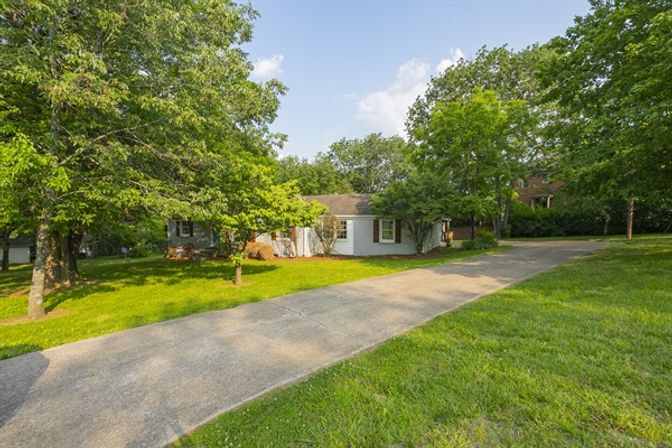 Tree-lined suburban ranch-style single-story home with a long concrete driveway, shaded lush front yard and mature trees on a sunny day